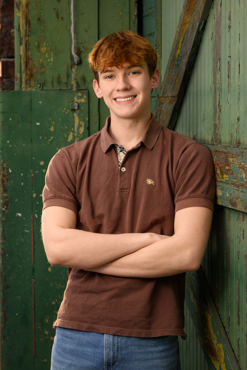 High school senior boy in brown polo leaning on a peeling green barn door, Spokane senior portrait