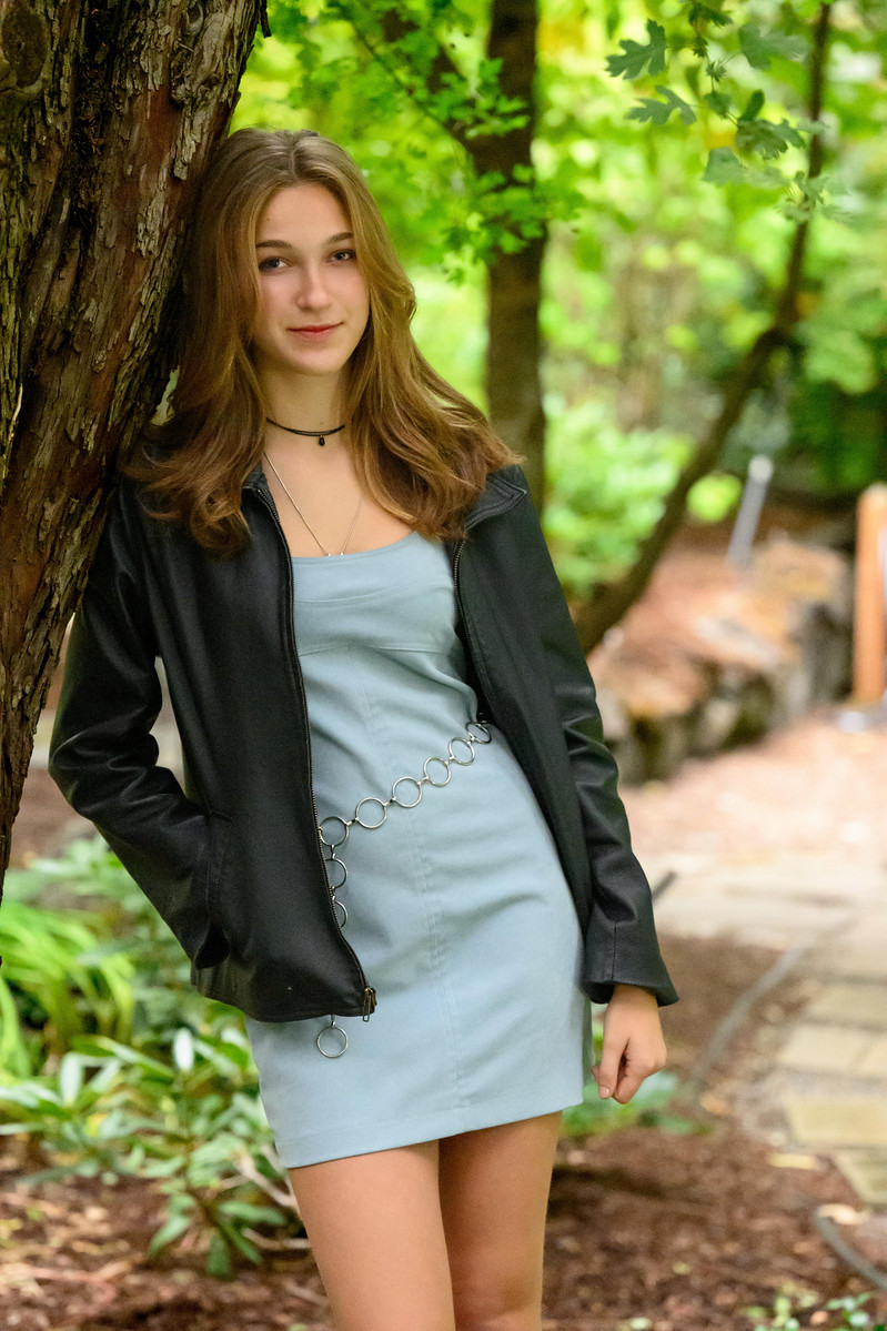 High school senior girl leaning against a tree in a lush green forest, wearing a light blue mini dress and black leather jacket — senior portrait photography in Spokane, WA